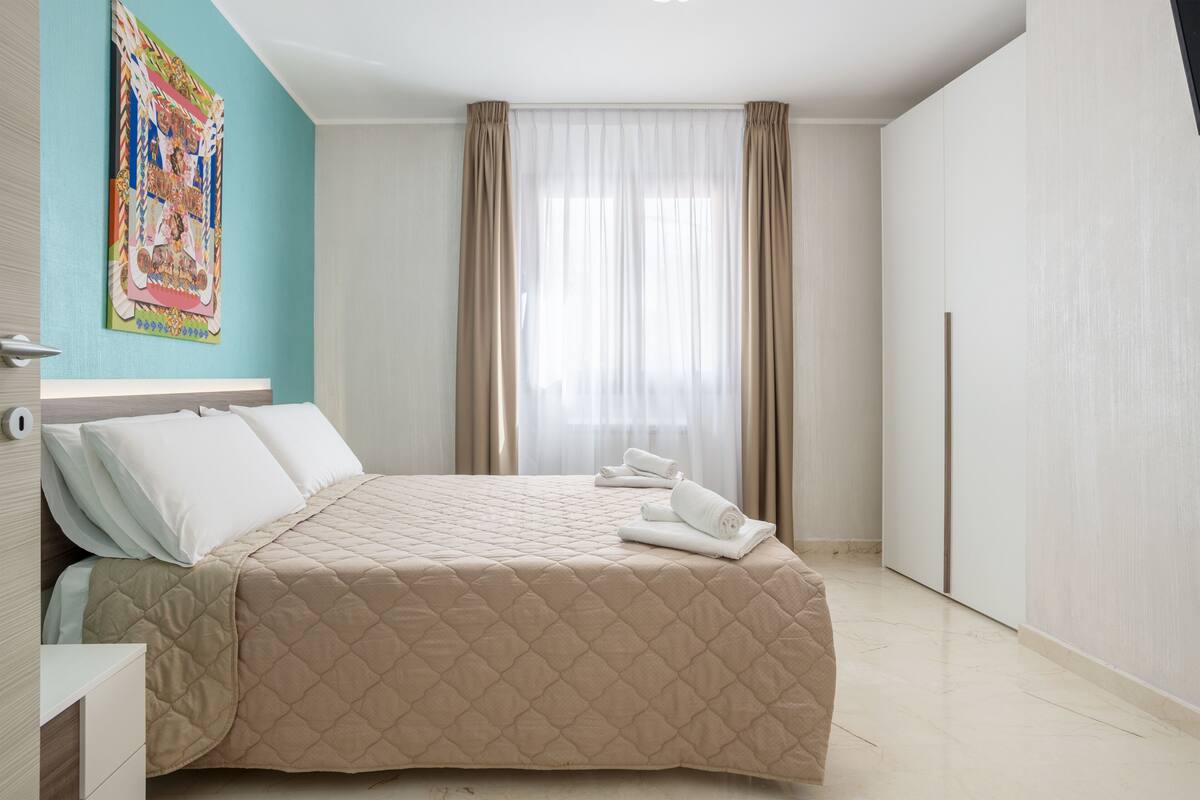 A modern hotel room with a large bed, a nightstand, a window with sheer curtains, and a colorful artwork on the wall.