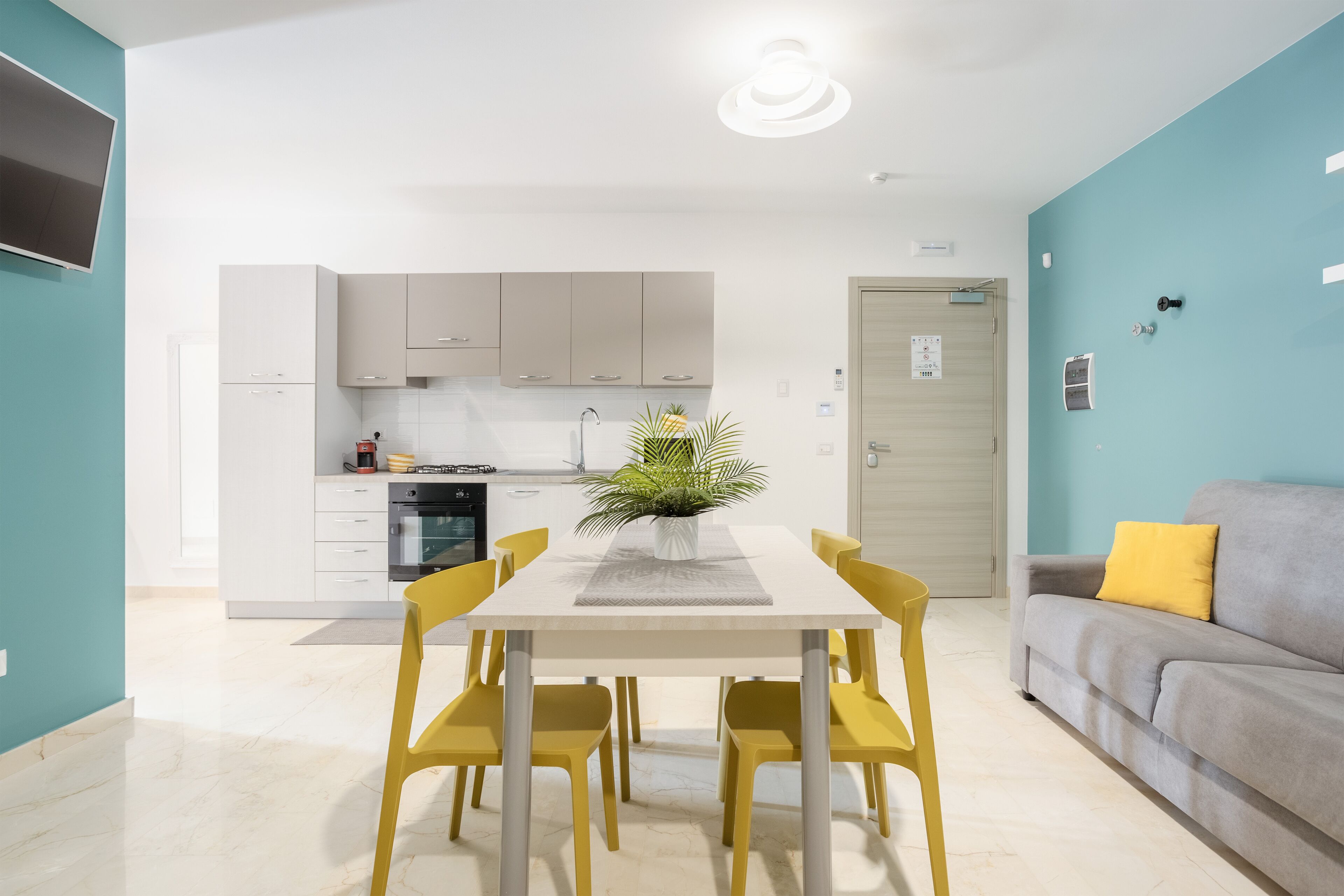 A modern kitchen with white cabinets, a dining table with yellow chairs, and a grey sofa with a yellow pillow.