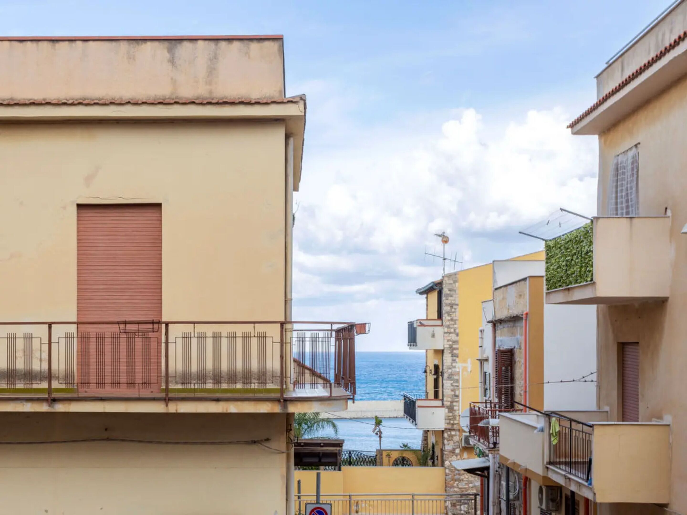 A coastal building with a red shutter, a balcony with metal railings, and a view of the sea.