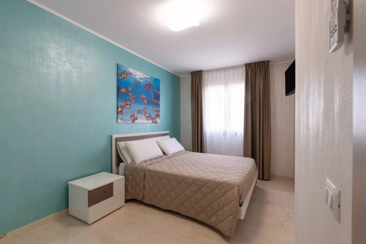 A bedroom with a bed, a nightstand, a painting on the wall, and a window with curtains.