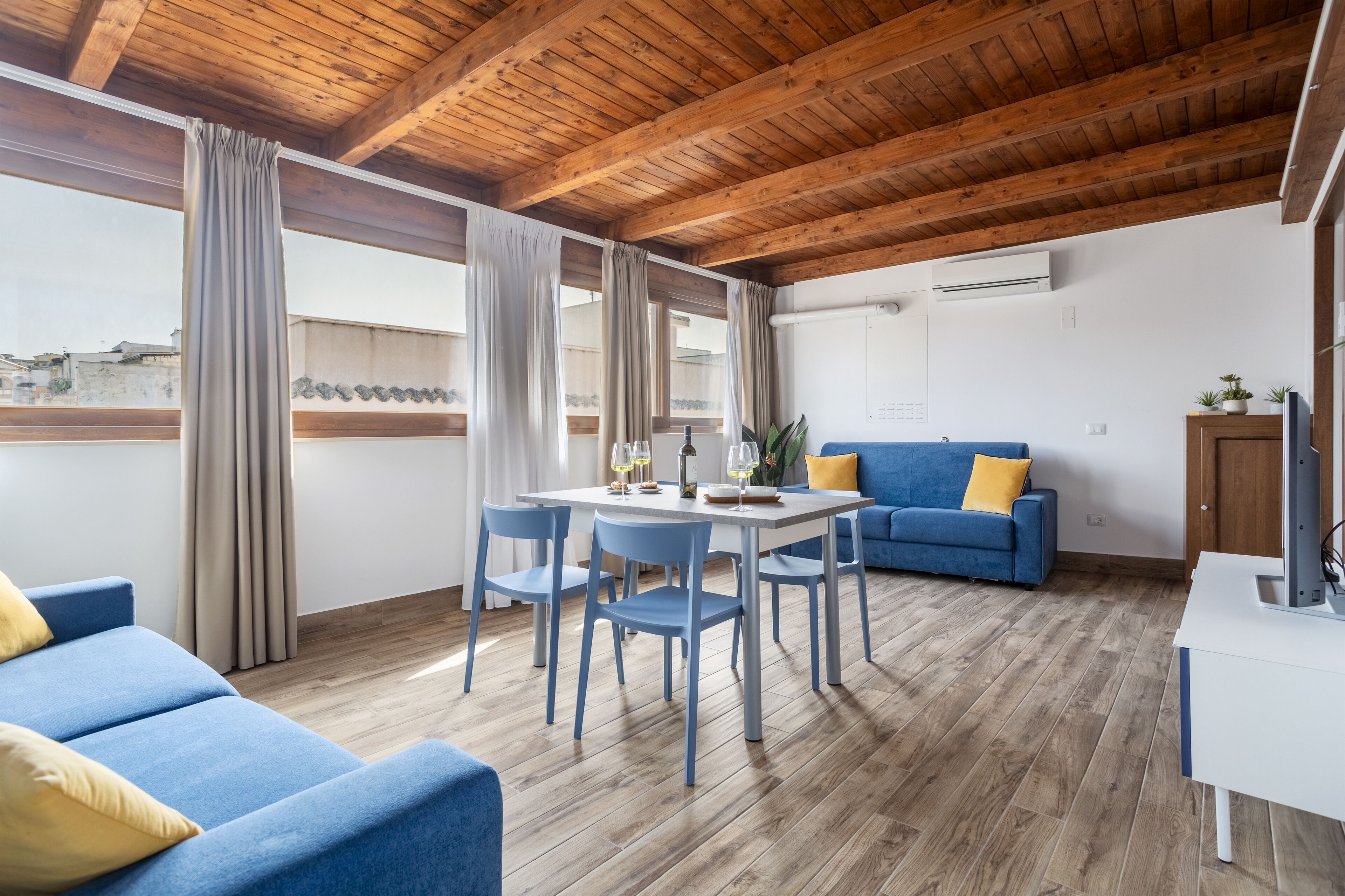 A modern living room with a wooden ceiling, a dining table with chairs, a blue sofa, and a small TV on a white table.