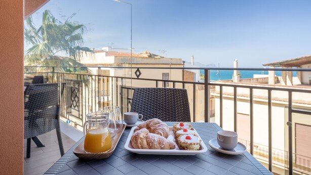 A balcony with a table set for breakfast, including croissants and a glass of orange juice, overlooking a coastal view. - Azzurra Apartments (Balestrate)