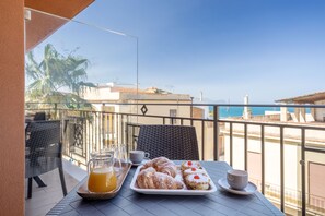 A balcony with a table set for breakfast, including croissants and a glass of orange juice, overlooking a coastal view. - Azzurra Apartments (Balestrate)