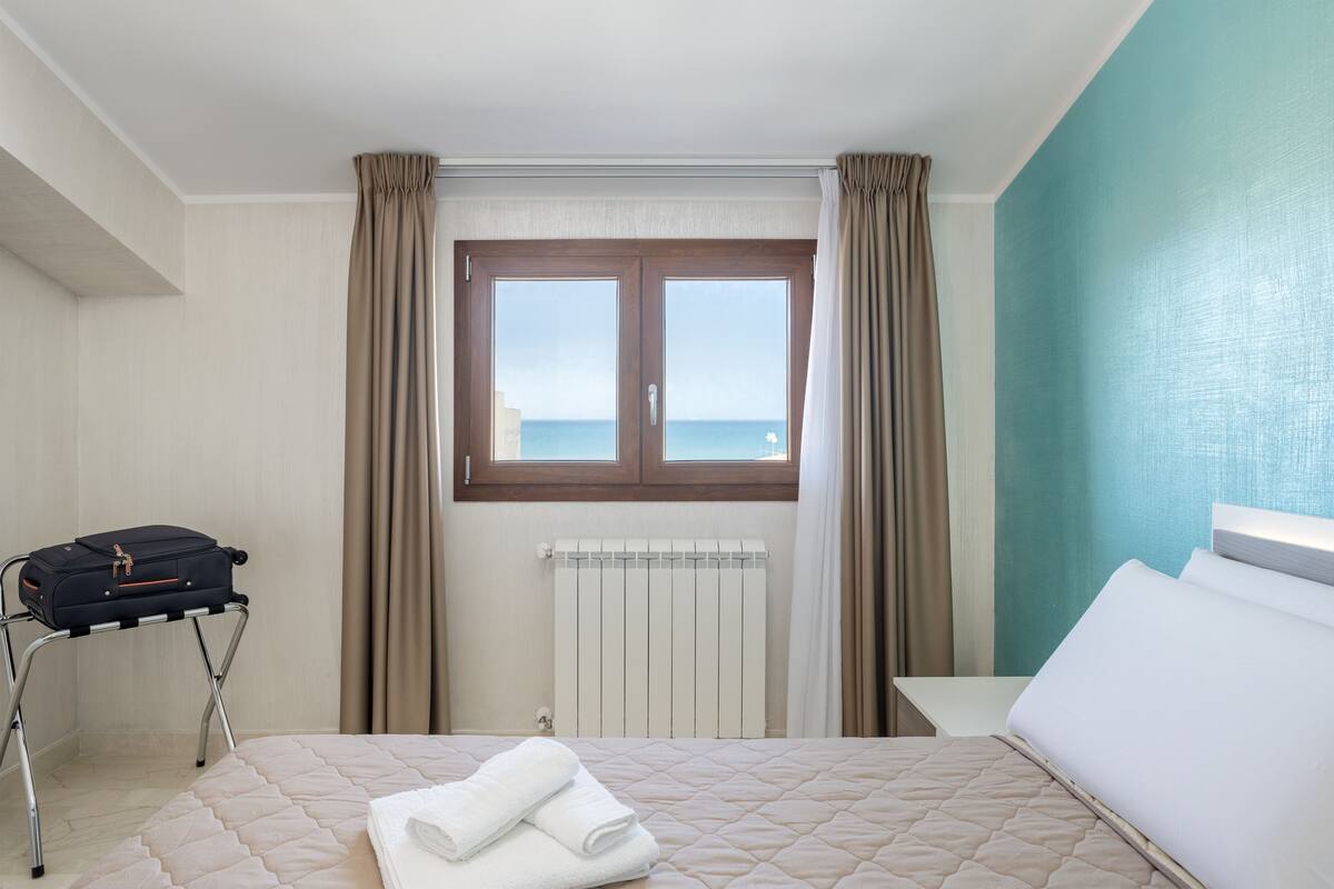 A hotel room with a bed, a suitcase on a trolley, a radiator, and a window with curtains.