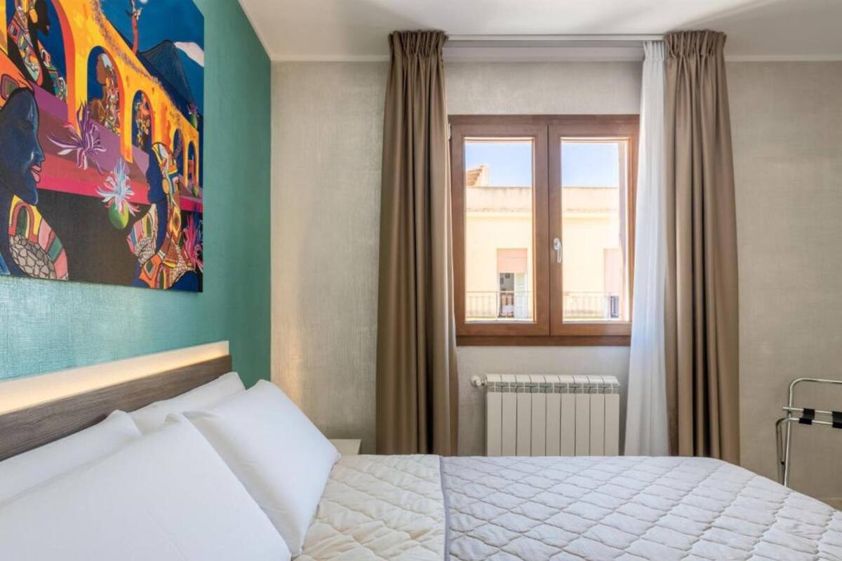 A hotel room with a bed, a window with curtains, a radiator, and a colorful painting on the wall.