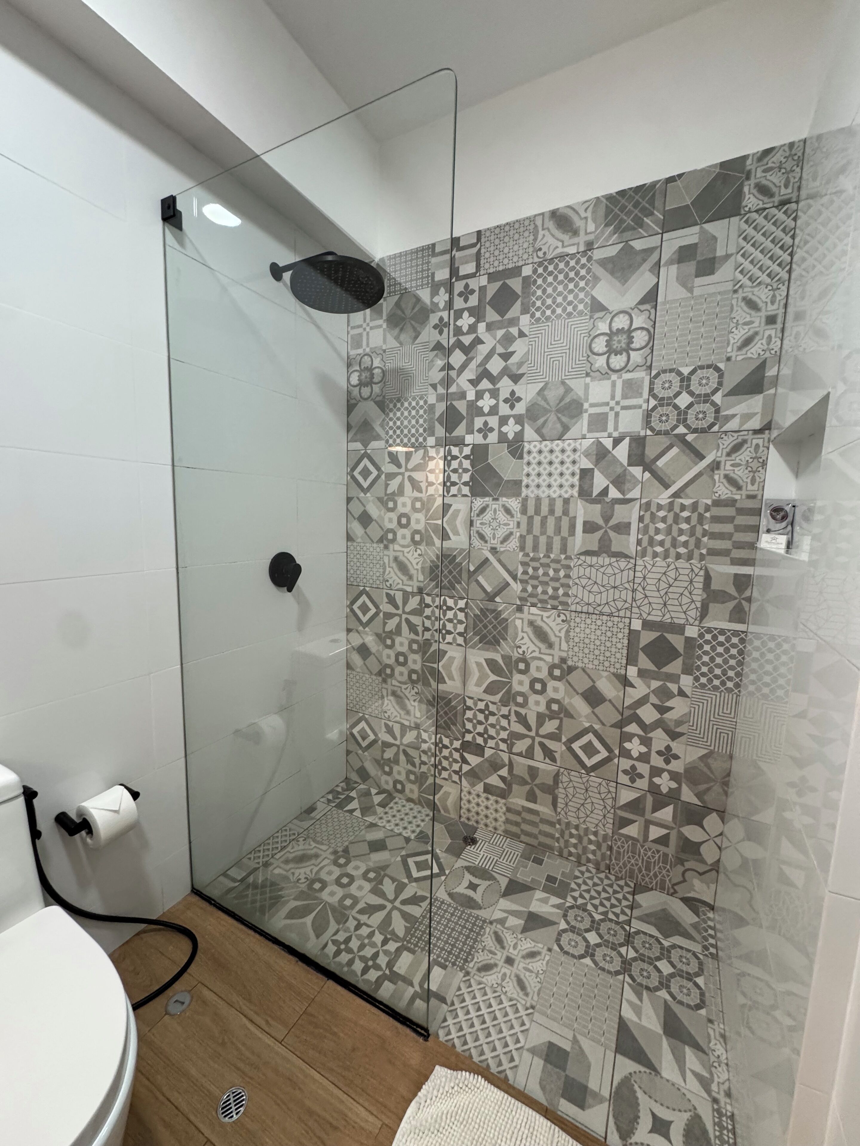 A shower with a patterned tile wall, a glass door, and a toilet with a visible pipe.