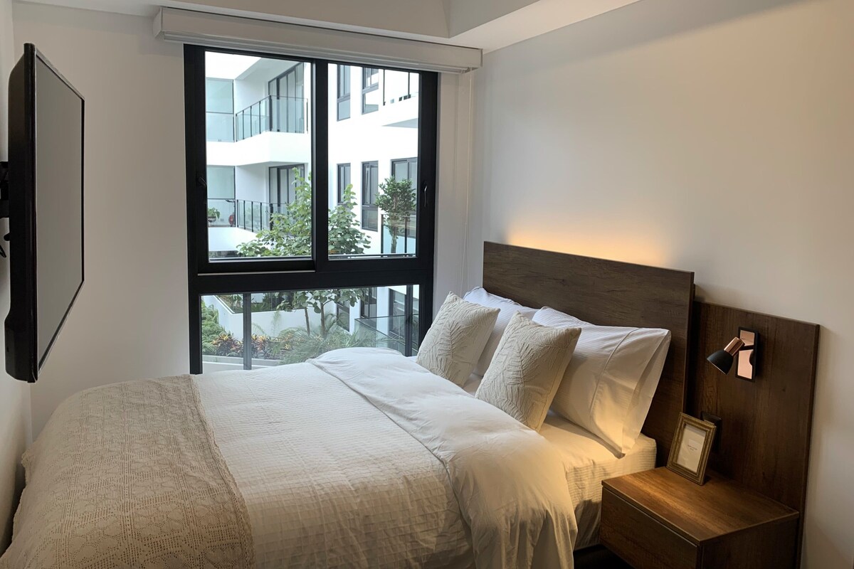 A modern hotel room with a large window, a neatly made bed, and a wooden nightstand with a lamp.