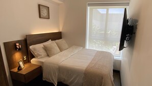 A neatly made bed with a wooden headboard, a bedside table with a lamp, and a window with a view of greenery. -  Aparts4Days Executive Apartment San Isidro Business (Lima)
