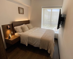 Una cama bien hecha con cabecera de madera, una mesita de noche con lámpara y una ventana con vistas a zonas verdes.