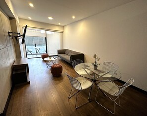 A modern living room with a glass dining table, wireframe chairs, a grey sofa, and a small orange ottoman. -  Aparts4Days Executive Apartment San Isidro Business (Lima)