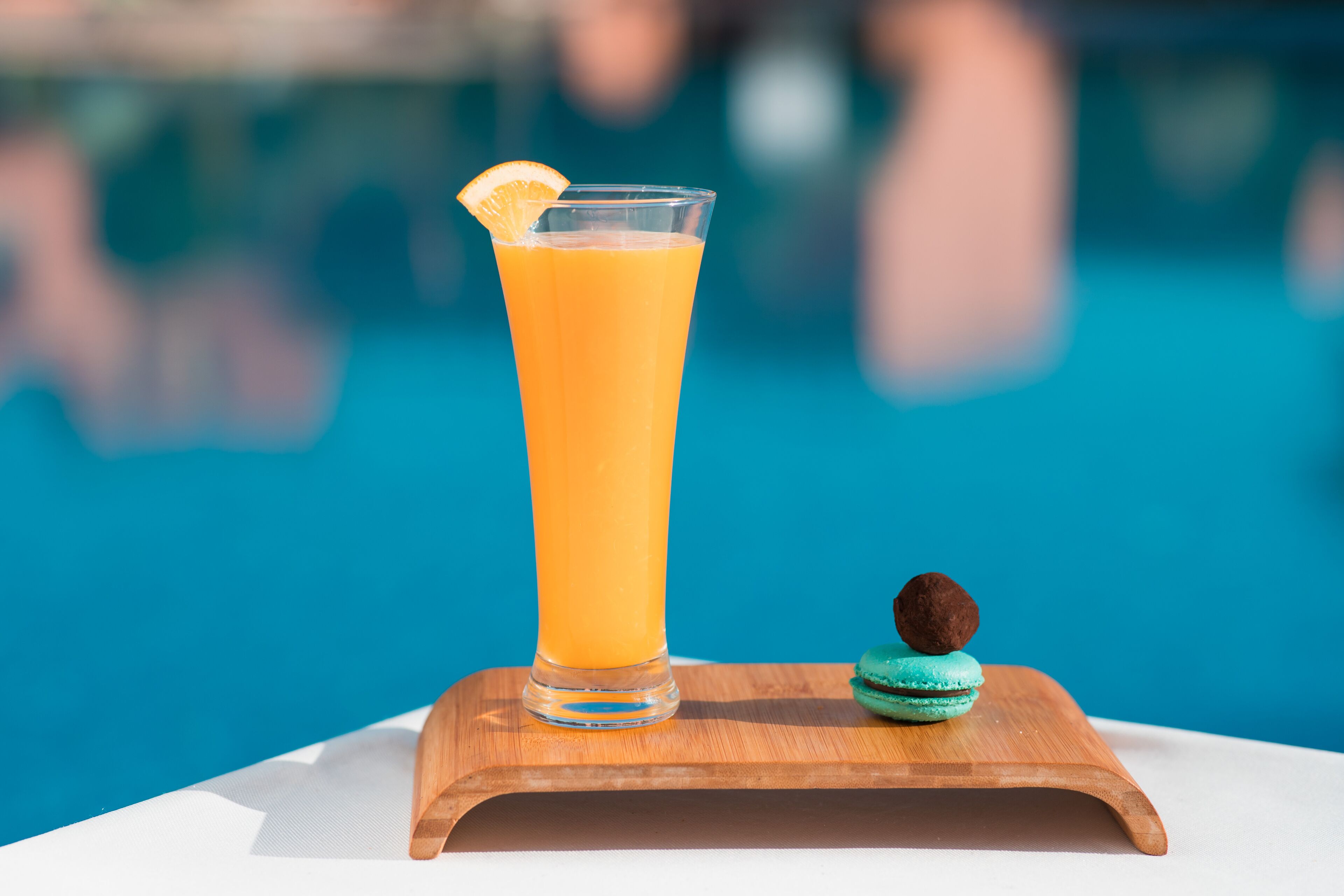Un vaso alto de jugo de naranja con una rodaja de naranja sobre una bandeja de madera junto a una piscina.
