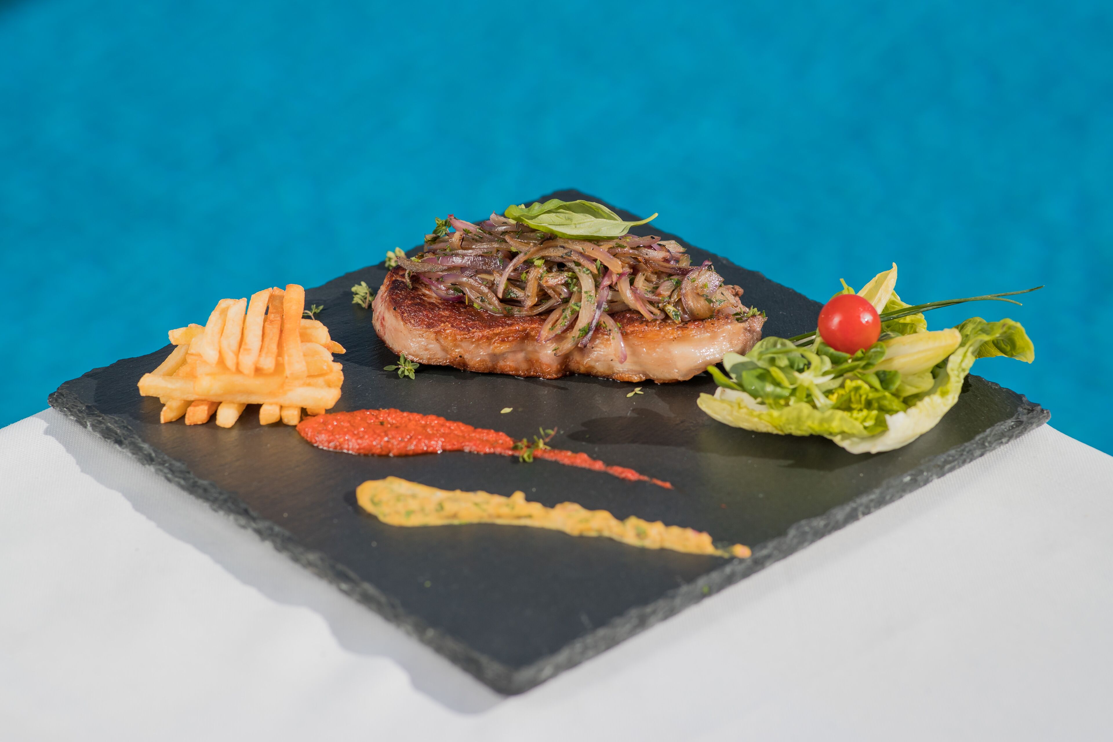 A black slate serving board with a grilled meat, fries, a red chili, a small salad, and a sauce.