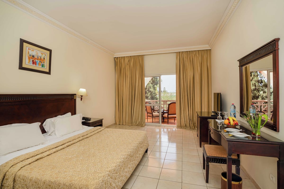 A hotel room with a bed, a desk with a mirror, a chair, a dining table with chairs, and a balcony with curtains.