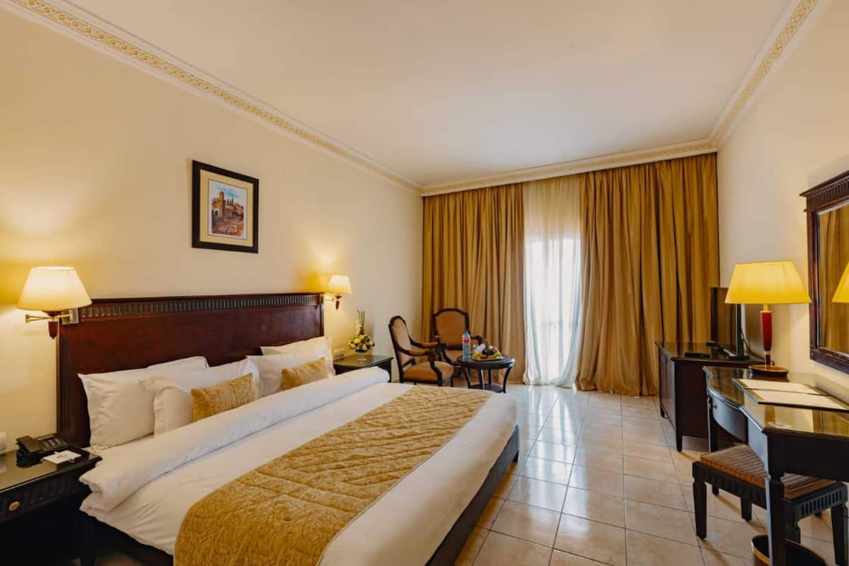 A hotel room with a large bed, two bedside tables with lamps, a desk with a chair, and a large window with curtains.