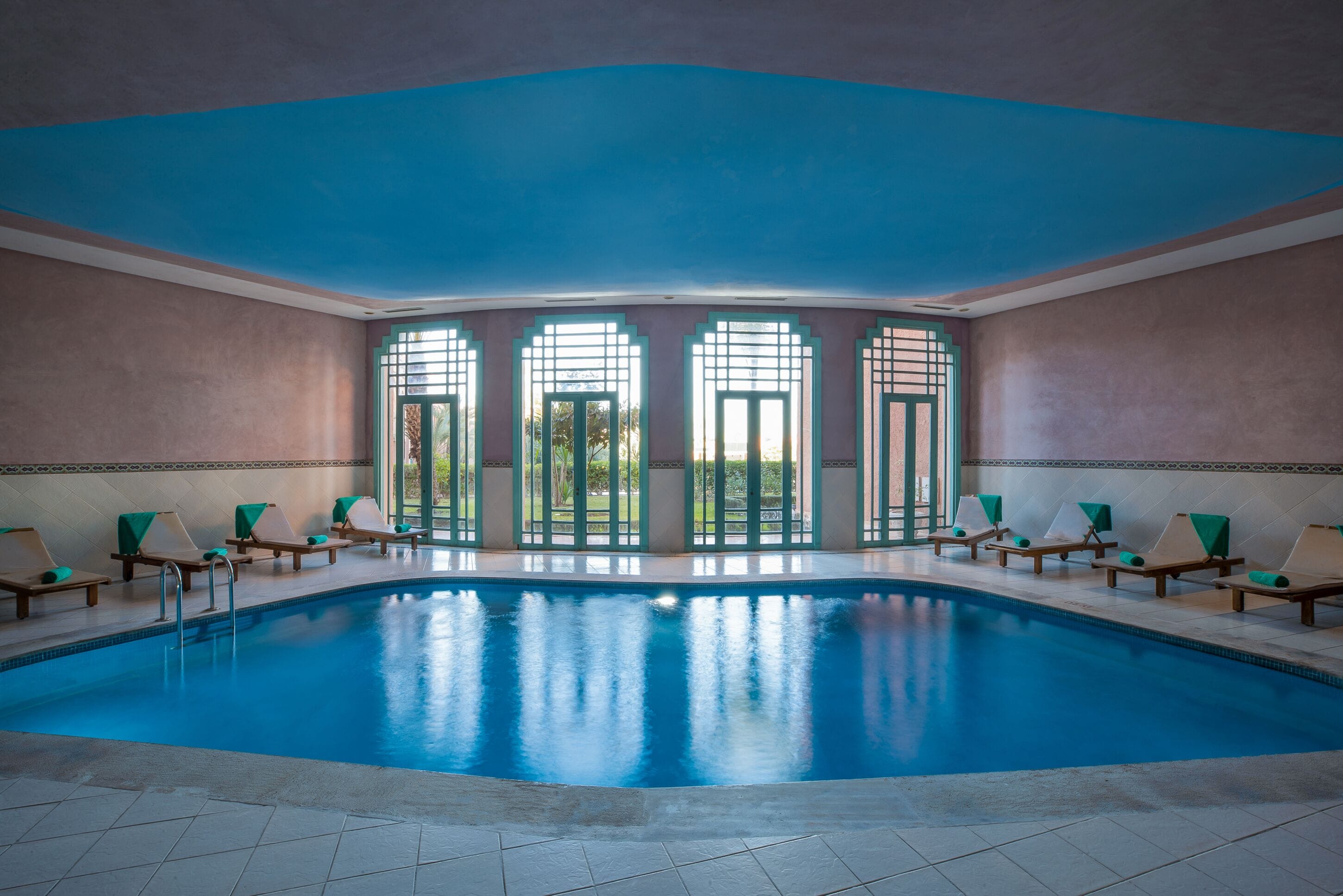 A large indoor swimming pool with a blue ceiling, surrounded by lounge chairs and large windows.