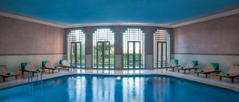 A large indoor swimming pool with a blue ceiling, surrounded by lounge chairs and large windows.