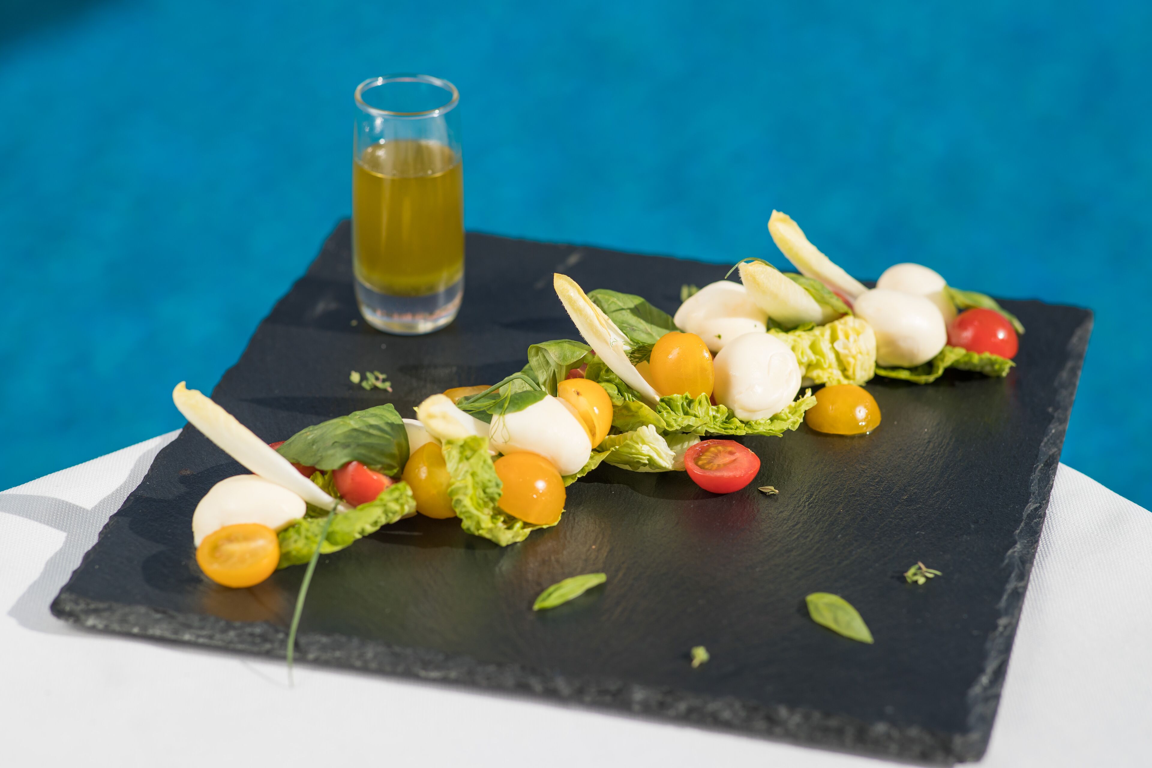 A black slate serving board with a salad consisting of lettuce, cherry tomatoes, mozzarella balls, and a glass of yellow liquid, possibly olive oil, on a blue background.