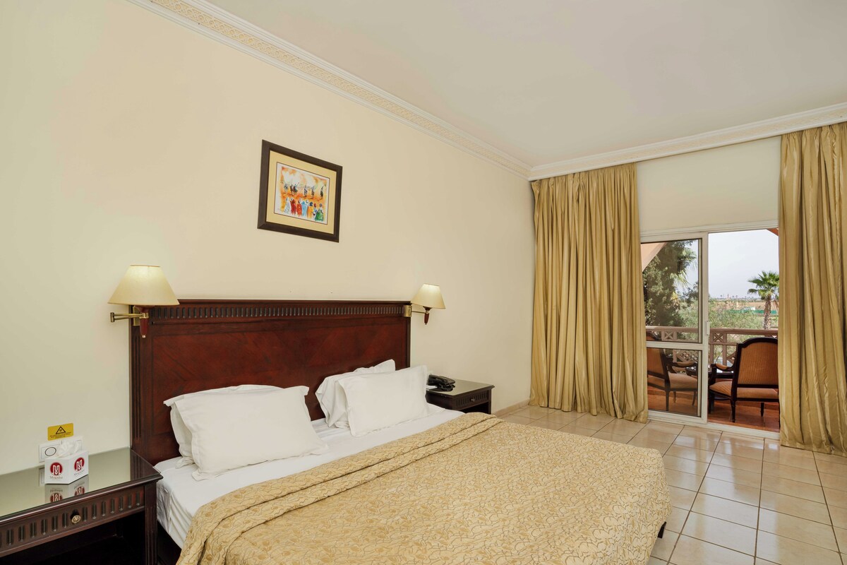 A hotel room with a bed, nightstand, lamp, and a view of the outdoors through a sliding door.