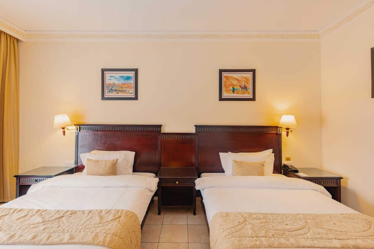 A hotel room with two beds, each with a wooden headboard and bedside lamps. There are framed pictures on the wall above the beds.