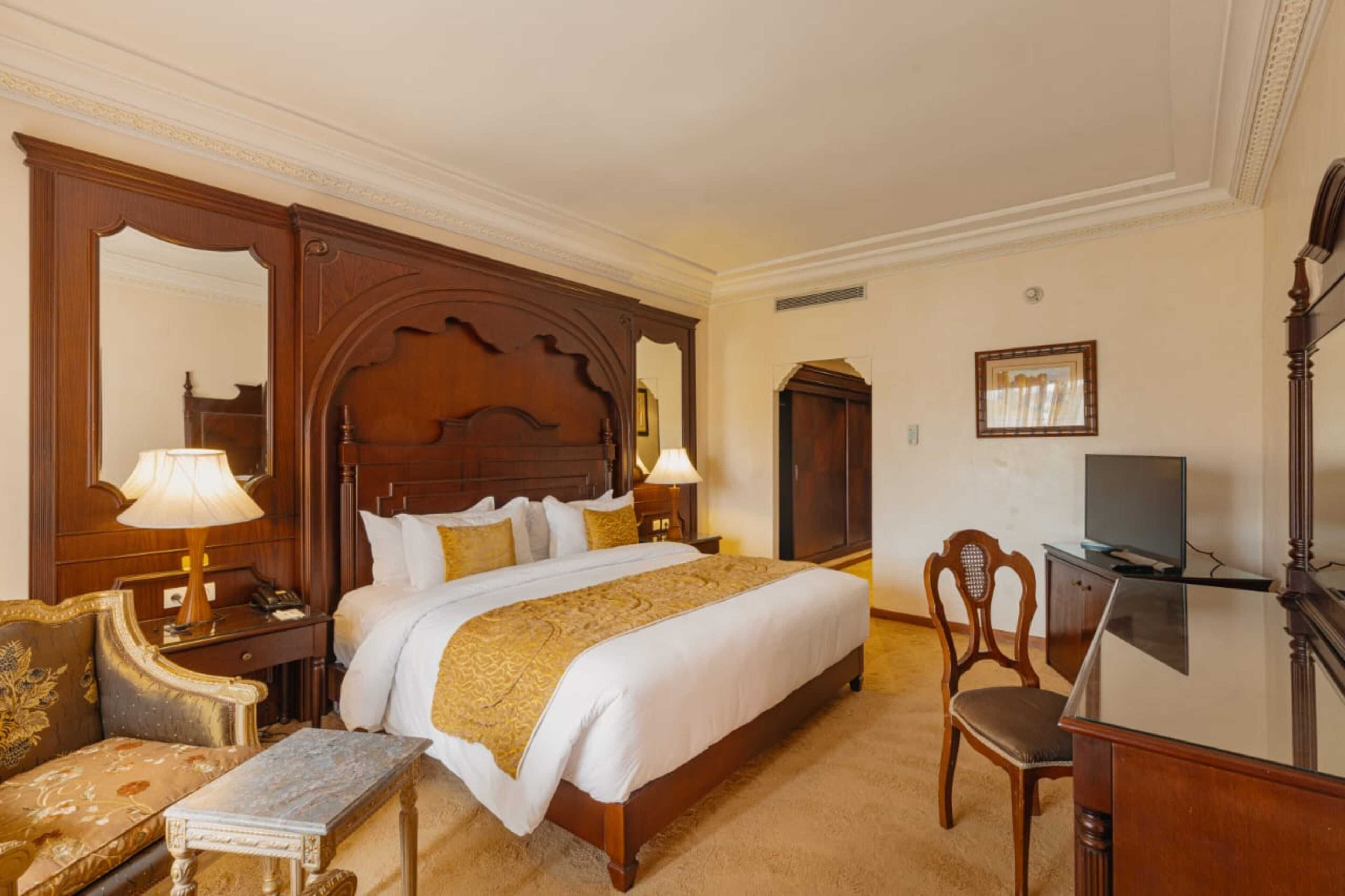 A hotel room with a large bed, wooden headboard, a desk with a television, a chair, and a small table.
