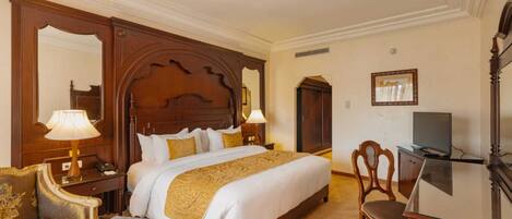 A hotel room with a large bed, wooden headboard, a desk with a television, a chair, and a small table.