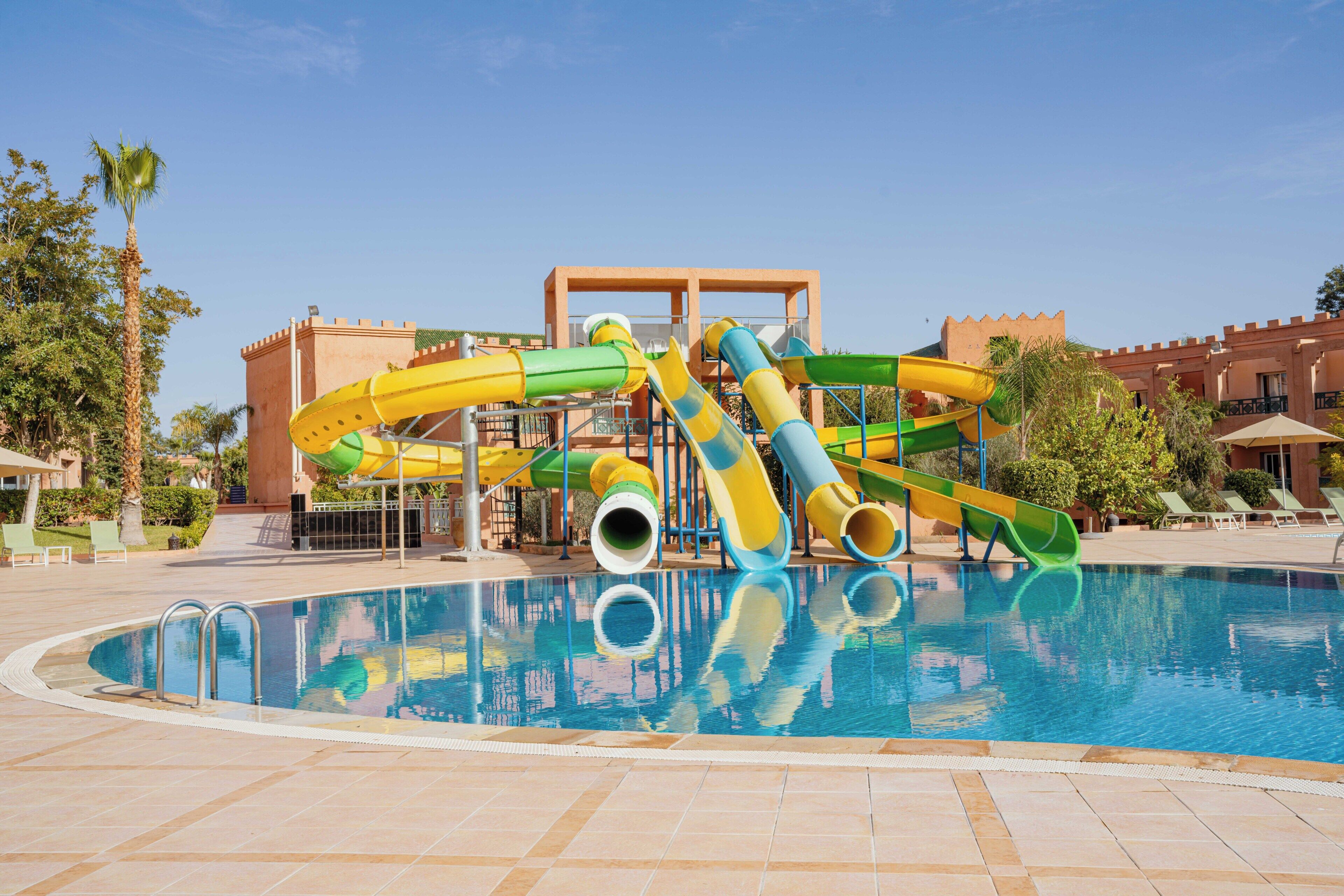 Una piscina con uno scivolo acquatico, palme e un edificio con facciata a castello.
