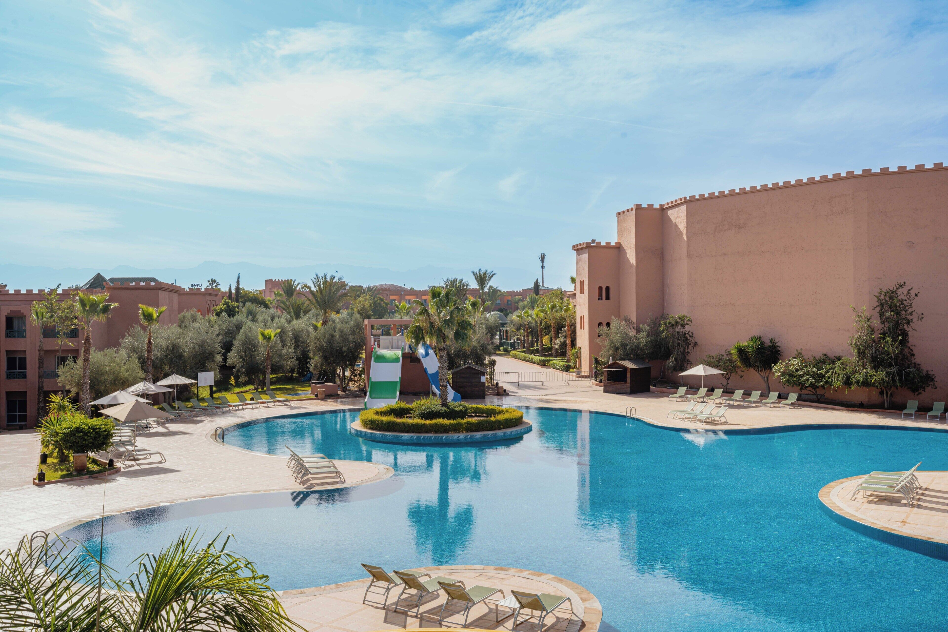 A swimming pool with lounge chairs and umbrellas, surrounded by palm trees and a castle-like building.