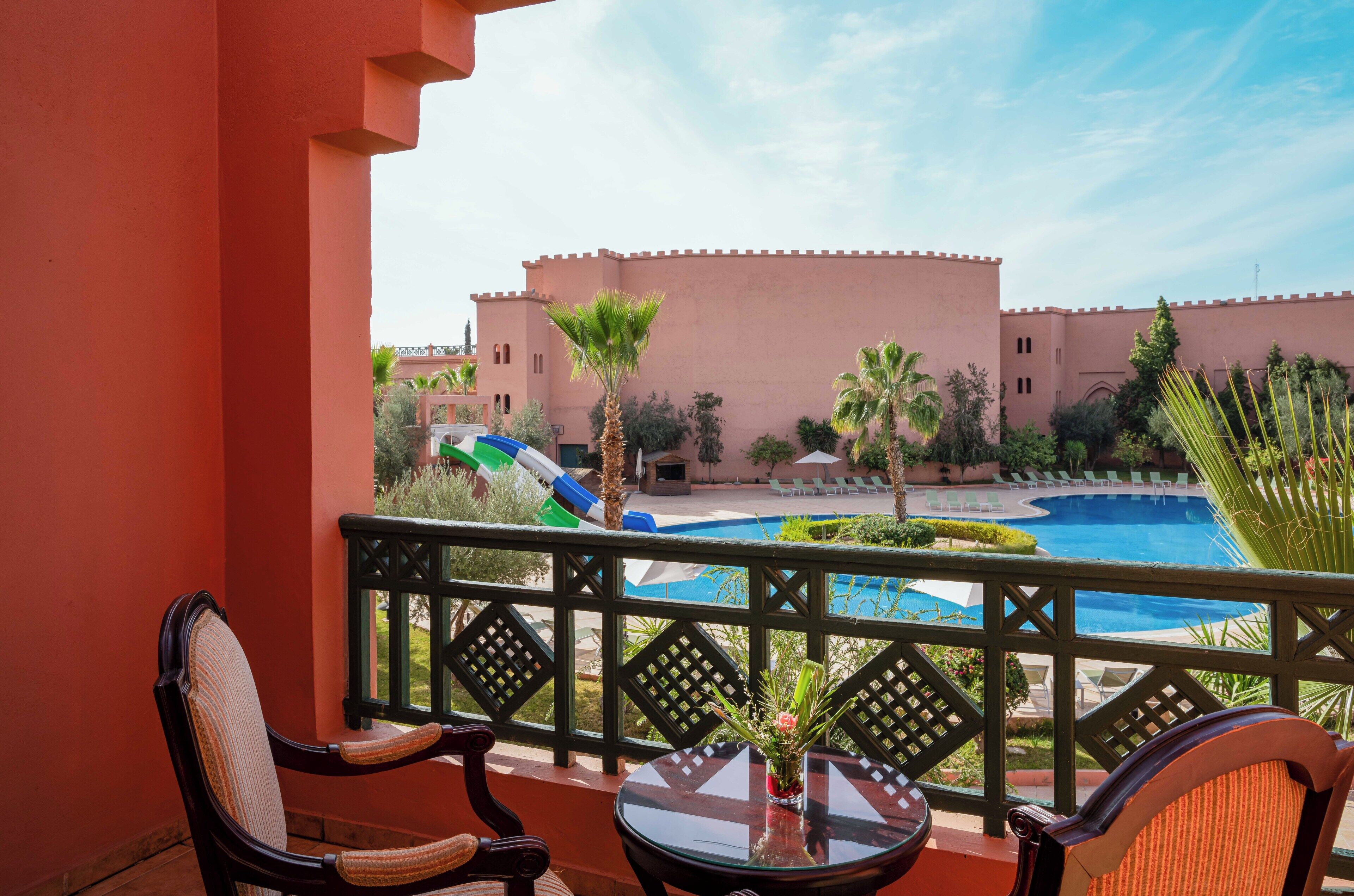 A balcony with a view of a swimming pool, palm trees, and a building with a pink facade.