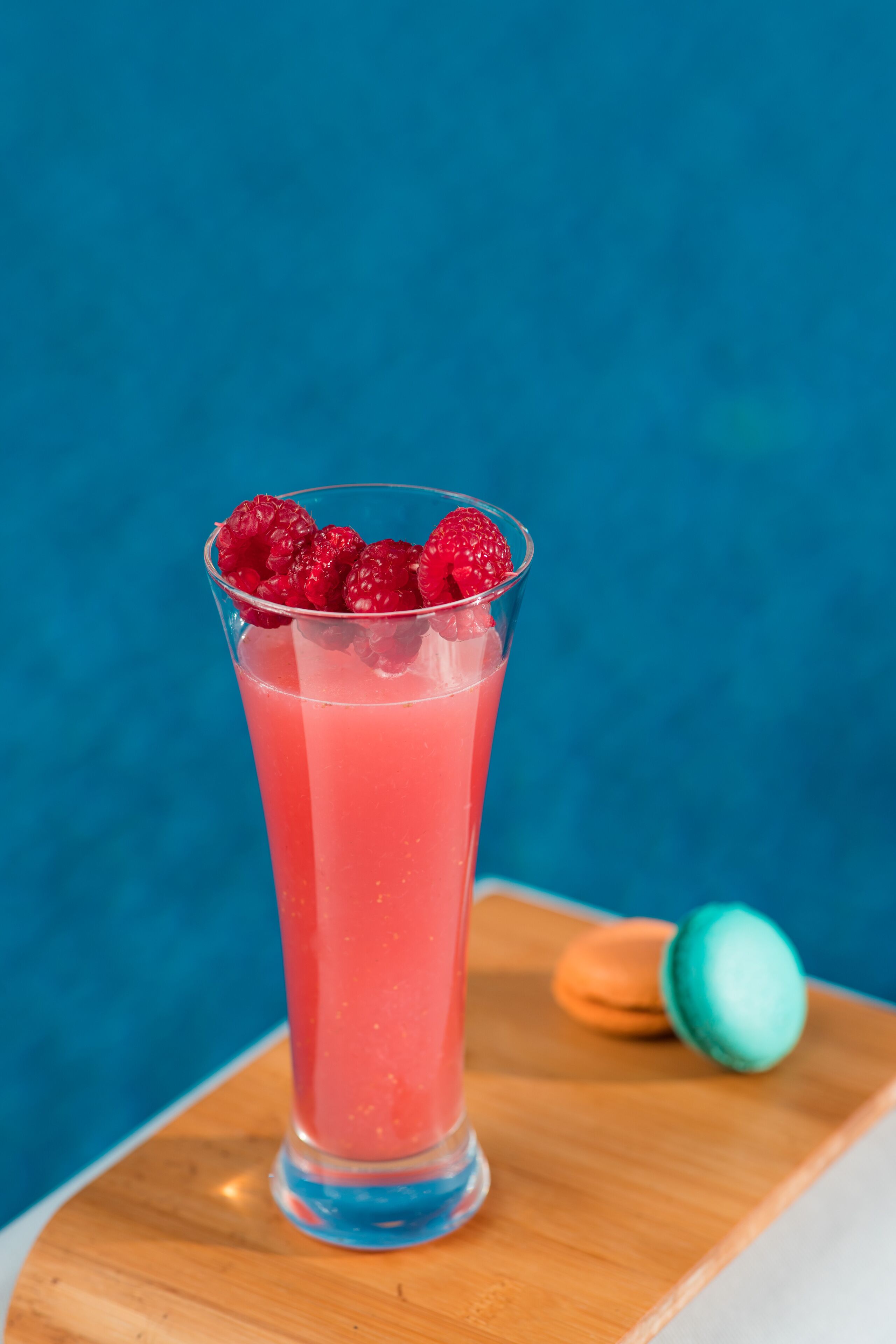Ein hoher Schluck mit einem rosa Getränk und Himbeeren, auf einer Holzoberfläche mit Macarons im Hintergrund platziert.