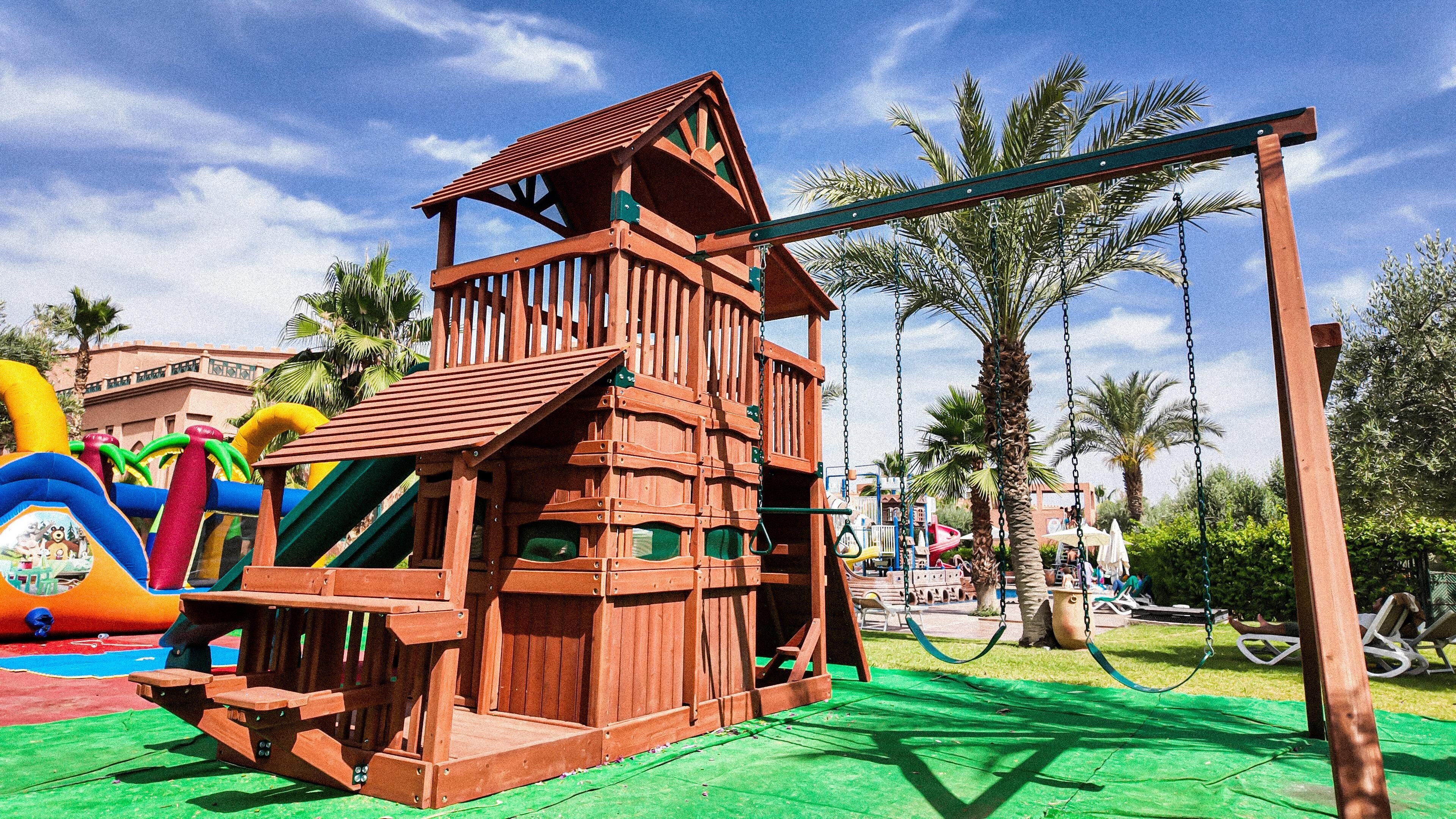 A wooden play structure with a slide and swings, surrounded by palm trees and a clear sky.
