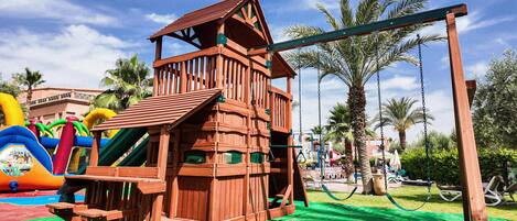 A wooden play structure with a slide and swings, surrounded by palm trees and a clear sky.