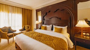 Habitación de hotel con una cama grande, cabecera de madera, zona de estar con una silla y mesita de noche con lámpara.