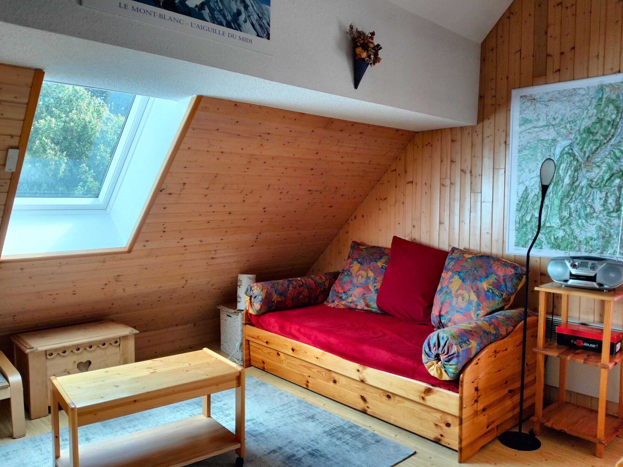Ein gemütliches Schlafzimmer im Loft-Stil mit einer schrägen Holzwand, einem roten Schlafsofa mit bunten Kissen, einem kleinen Holztisch, einem Radio auf einer Holzablage und einer Karte an der Wand.