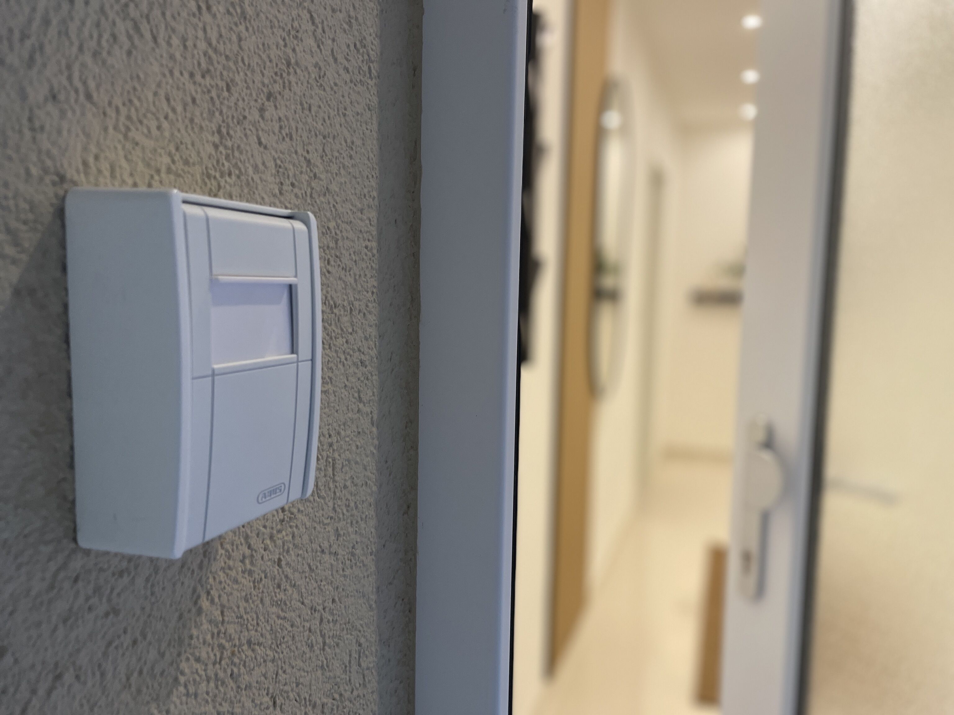 Une boîte électrique blanche est fixée à un mur texturé, et mène à un couloir où se trouvent une porte et un luminaire.