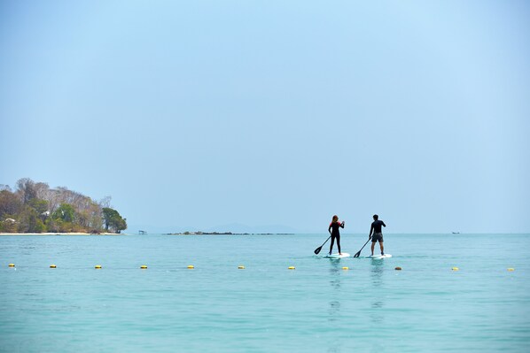 Two people paddleboarding on a calm sea with buoys, a beach with trees and buildings in the background, and a clear sky. - Villa Escape Burasari (Maprao Island)