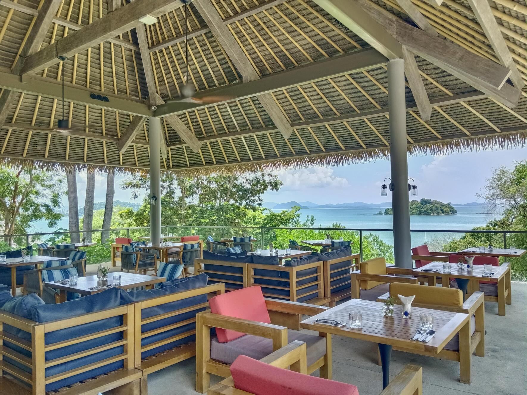 An outdoor dining area with wooden tables and benches, a thatched roof, and a view of the sea.
