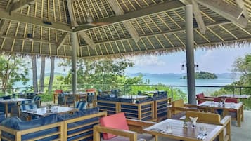 An outdoor dining area with wooden tables and benches, a thatched roof, and a view of the sea.