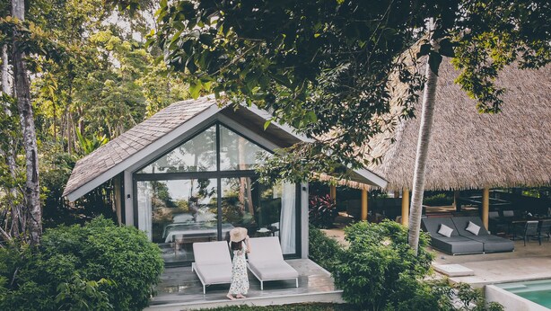 Una villa moderna con fachada de vidrio, una estructura de techo de paja y una zona de piscina.