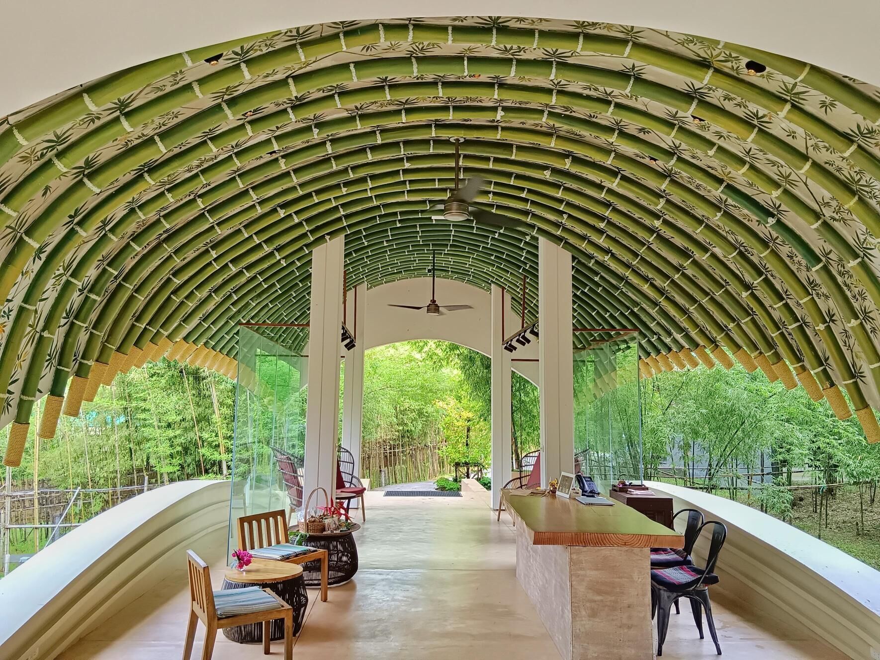 Un espacio de estar al aire libre cubierto, con una estructura de arco de bambú, barandas de vidrio y una mesa central con sillas.