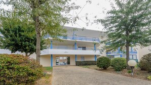 A two-story building with a blue balcony, a gravel driveway, and surrounding greenery.