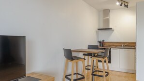 A modern kitchen with a wooden countertop, a built-in oven, and a stainless steel sink. There are three bar stools with black seats and wooden legs. A flat-screen TV is mounted on a wooden stand. The room has white walls and a wooden floor.