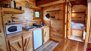 Una cabaña de madera compacta con kitchenette, un ventanal pequeño y una cama con colcha colorida.