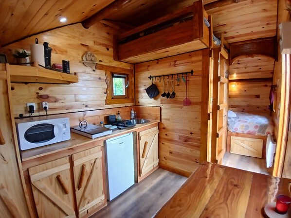 Eine gemütliche Holzhütte mit Küchenzeile, kleinem Fenster und einem Bett mit bunter Bettdecke.