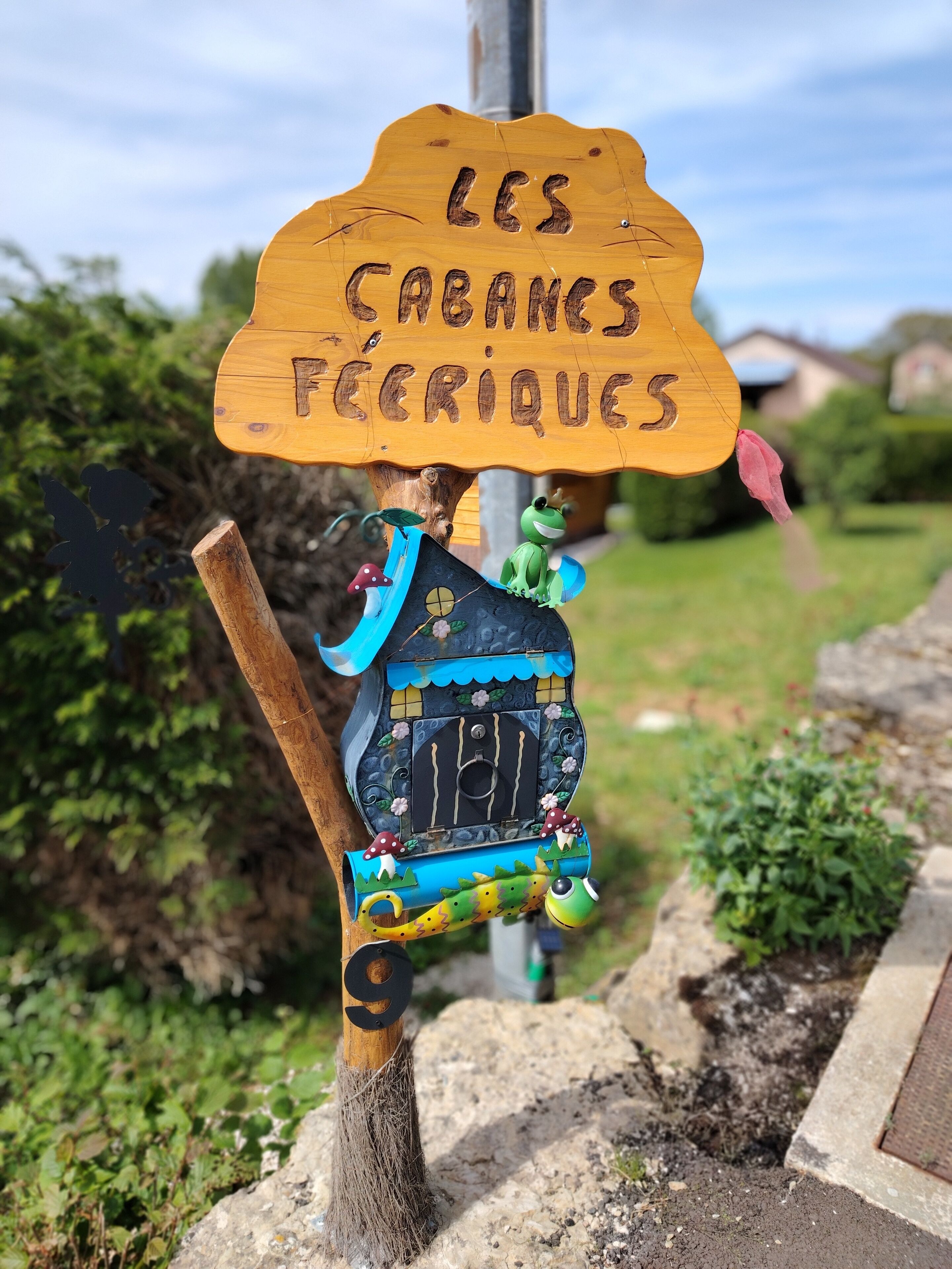 A wooden sign with the text 'LES CABANES FÉRIQUES' and a birdhouse with a frog figure and decorative elements.