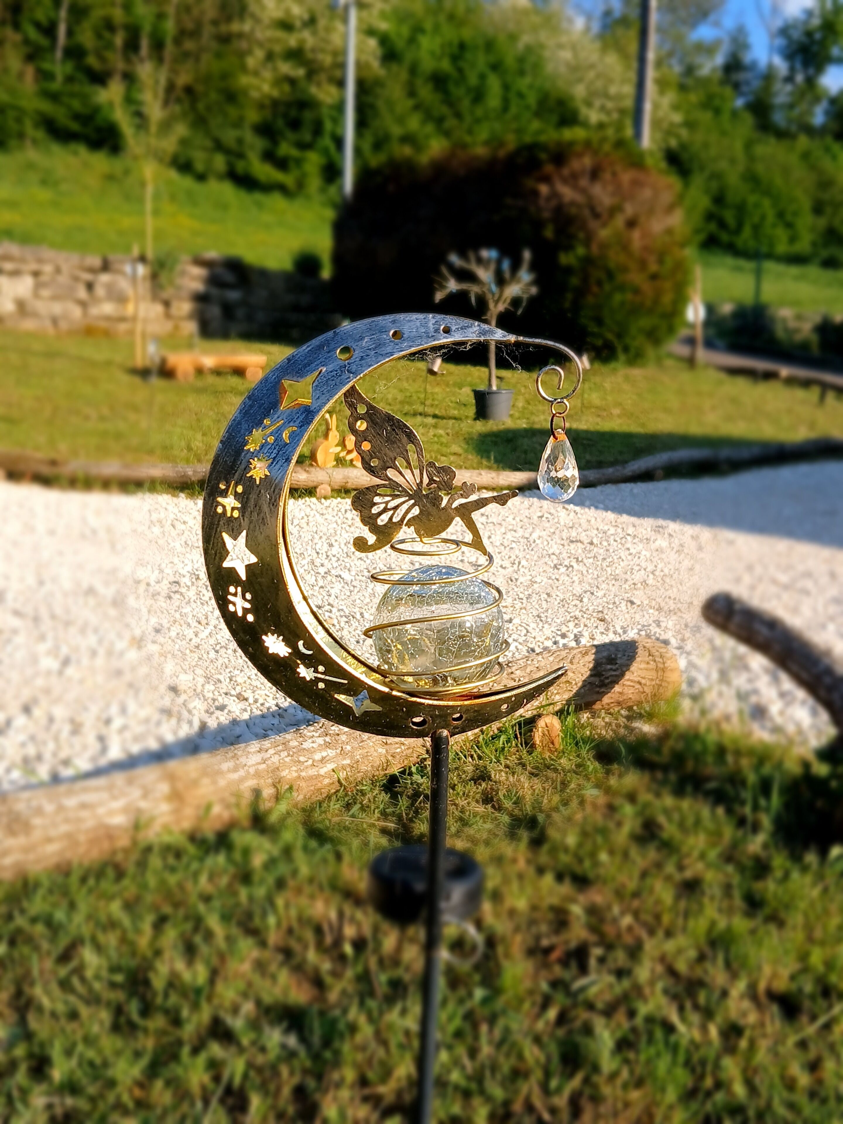 A decorative metal crescent with a glass orb and a hanging ornament, set on a gravel path with grass and trees in the background.