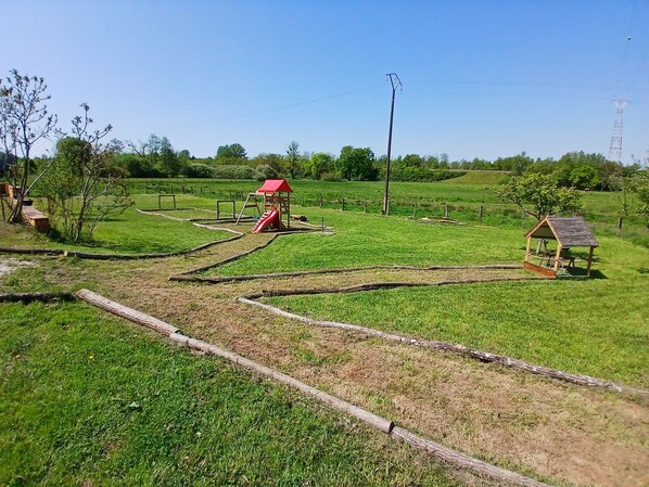Paisaje rural con un parque infantil, una pequeña construcción de madera y una zona cercada.
