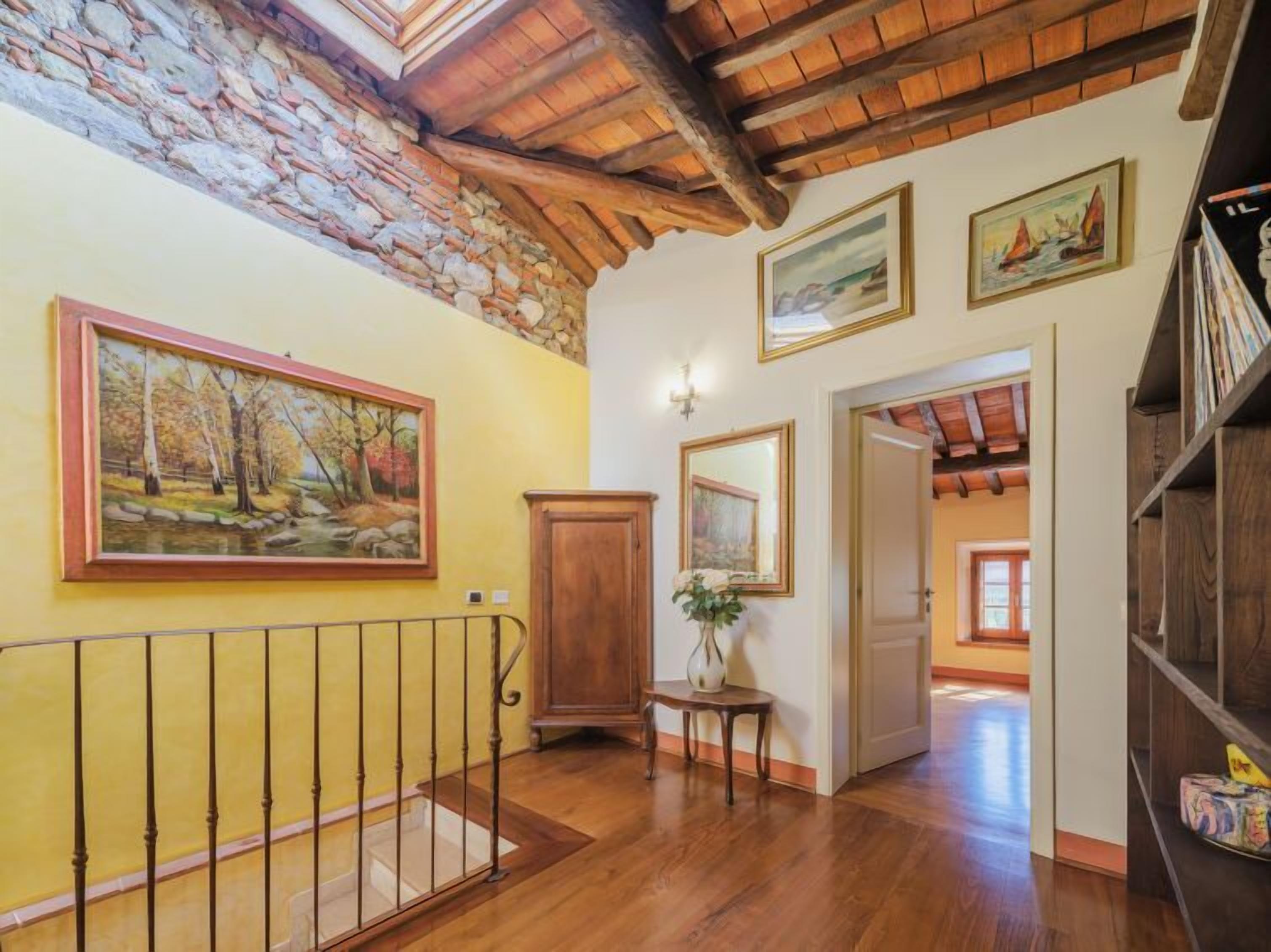 A spacious hallway with wooden flooring, a stone wall, and a wooden ceiling. There are paintings on the walls and a bookshelf on the right.