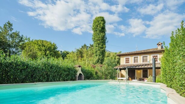 Uma piscina com água azul cristalina, rodeada por vegetação exuberante e um edifício com telhado em azulejos.