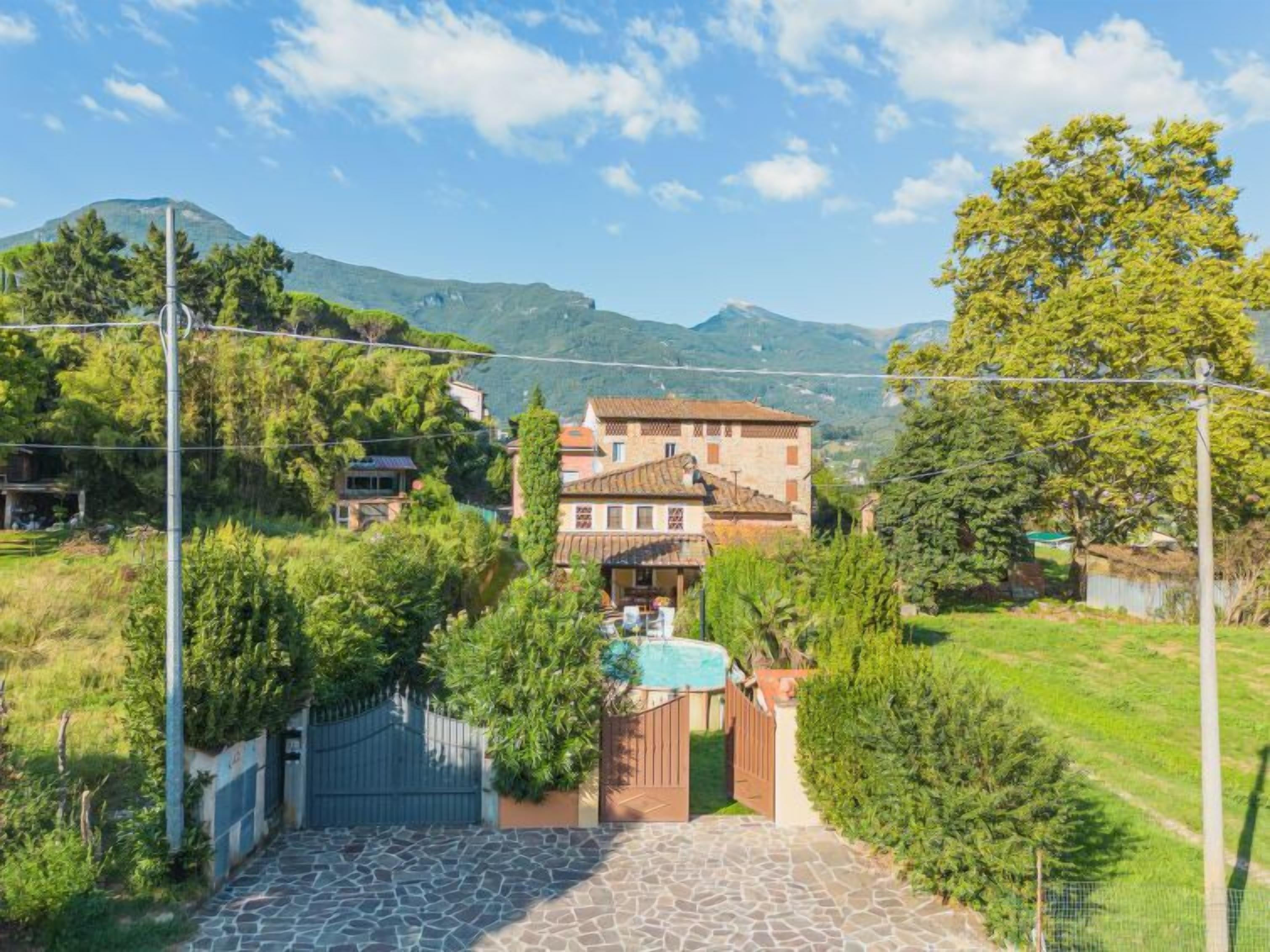 A two-story stone house with a tiled roof, surrounded by lush greenery and a mountain in the background.