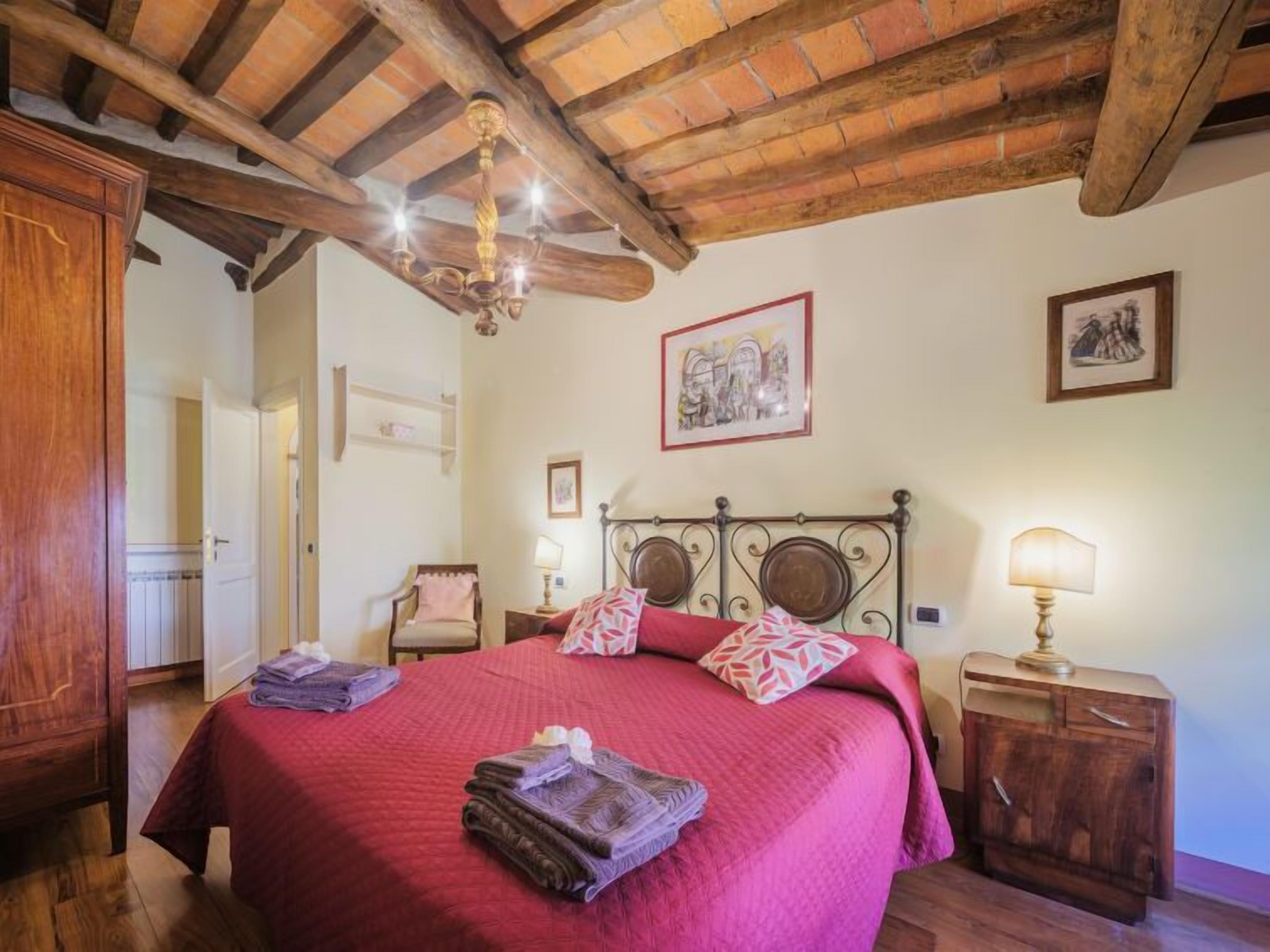 A bedroom with a wooden ceiling, a bed with red bedding, two bedside tables with lamps, and a wooden wardrobe.